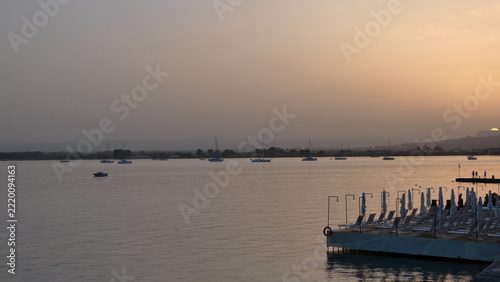 Calm bay at dusk with sailboats anchored under a hazy orange sky