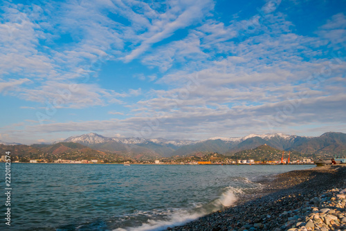 View of the blue sky with white clouds over the mountains in Batumi, Georgia. Waves of the Black Sea are crashing on the pebble beach - wide angle view