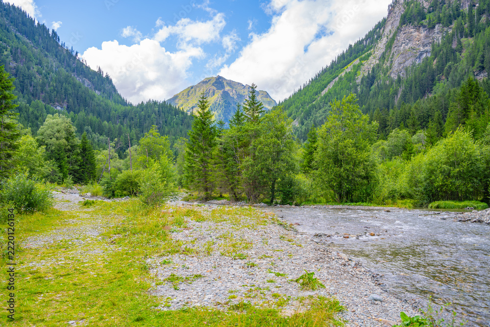 Naklejka premium In the Weisspriach Valley, tall trees line the riverbank. The sunlight shines on the mountains and clouds above. Visitors can explore the trails and enjoy the scenery in Austria.