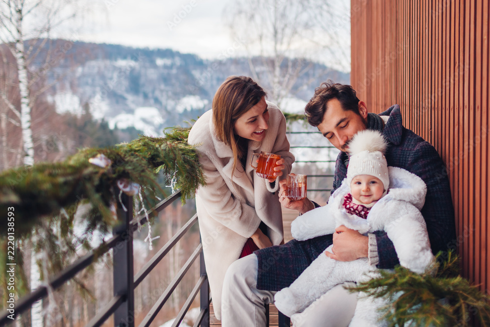 Naklejka premium Happy family drinking tea enjoying snowy mountains landscape from hotel balcony. Mom and dad having fun with toddler baby girl