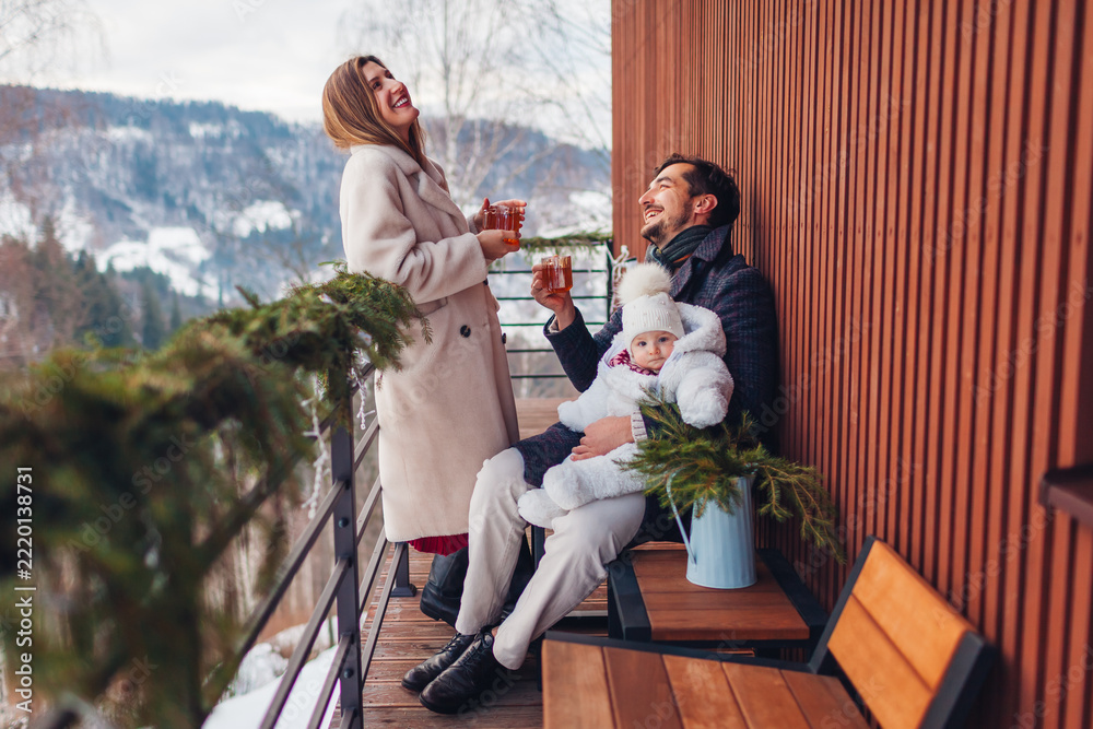 Fototapeta premium Happy family drinking tea enjoying snowy mountains landscape from hotel balcony. Mom and dad having fun with toddler baby girl