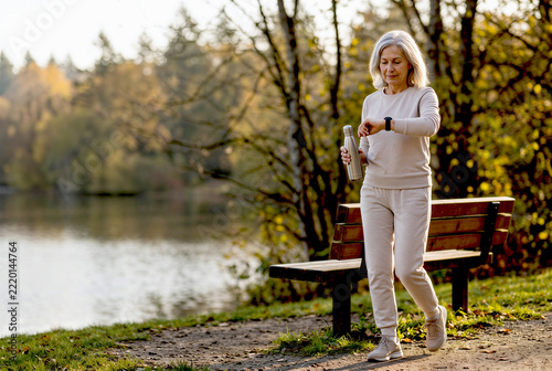 Silver-Haired Woman Walking Along Autumn Lakeside Path, Holding Water Bottle and Checking Smartwatch, Promoting Active Aging, Healthy Lifestyle and Outdoor Fitness with Copy Space
