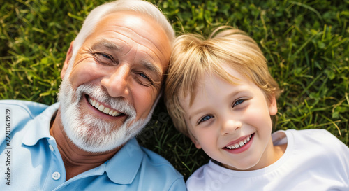 Top view close-up of smiling grandfather and grandson lying on green grass in sunlight, concept for family bonding, active aging and happy childhood
