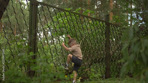Little Boy Climbs a Chain Link Fence in the Forest