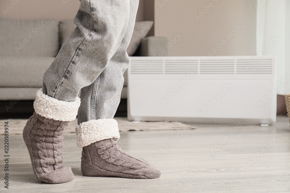 Fototapeta premium Cozy home comfort and winter warmth, person wearing soft knitted wool socks and jeans standing near a heater in a living room during cold weather
