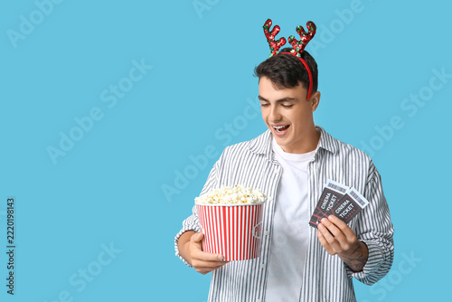 Young man in reindeer horns with cinema tickets and bucket of popcorn on blue...