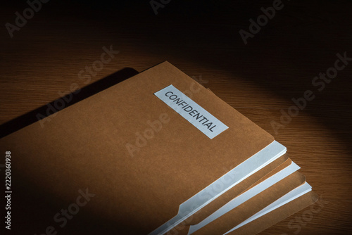 Stack of brown manila folders labeled CONFIDENTIAL on a wooden desk. Corporate secrecy, classified documents, legal privacy concept.