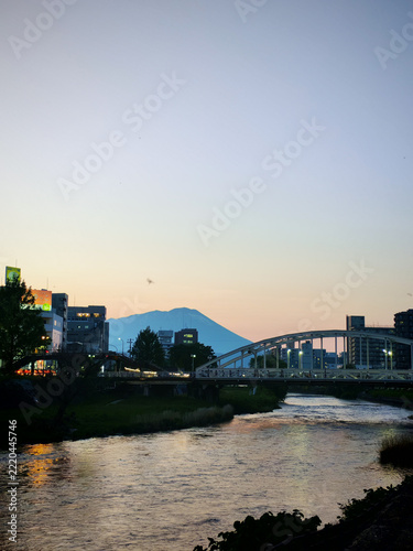 Serene evening view of cityscape with river flowing under bridge, silhouetted against pastel sky. Buildings line riverbank, and distant mountain adds to tranquil scene