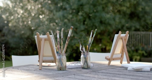 Camera is widening, shifting focus, revealing easels with brush jars on garden table to frame scene