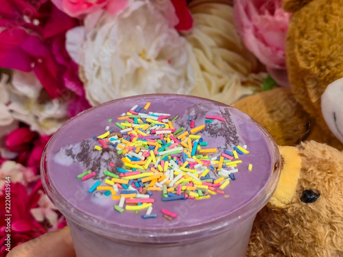 Fotografie Hand holding a cup of purple ube latte or lavender raf with colorful sprinkles, a teddy bear peeking from behind, flower wall of roses behind