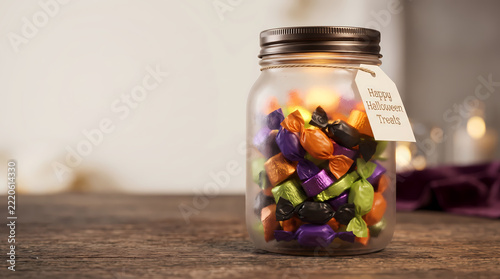 Glass jar filled with orange, purple, green and black wrapped Halloween candies with Happy Halloween Treats gift tag on rustic wooden table, copy space