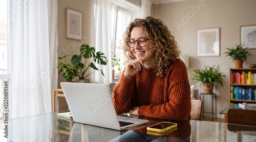 Happy young woman working from home on a laptop computer, sitting at a glass dining table in a bright modern living room with houseplants, wearing an orange sweater and glasses, holding a stylus pen