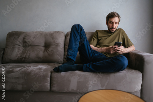 Man relaxing on a comfortable sofa, leaning back against the cushions and interacting with his mobile phone, browsing online content or sending messages in a casual setting