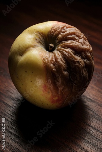 Close up of single rotten apple on dark wood table. Spoiled red, yellow fruit shows heavy decay, wrinkled skin. Decomposed organic food sits alone in shadow. Concept of waste, death, unhealthy eating