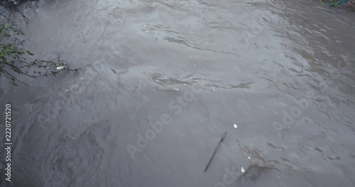 Camera looks down as a raging, sludge filled river surges through the narrow channel, sweeping along scattered debris, waste, and tree branches in its turbulent path