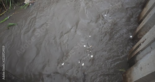 View captures a powerful and muddy small river, with the torrent of water rushing downstream and carrying debris through the swollen channel.