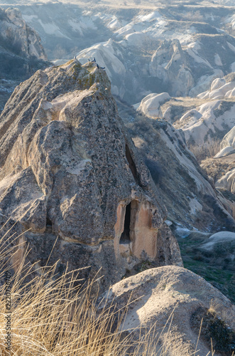 Fotografie Ancient rock-cut cave dwelling carved into a volcanic tuff formation overlooking a rugged valley in Cappadocia, Turkey, surrounded by distinctive eroded rock formations and sparse winter vegetation