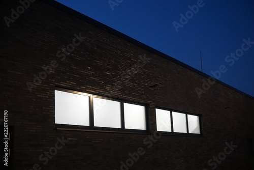 Moderne Architektur mit schwarzer Silhouette von Fassade einer großen Gebäudehalle, Flachdach und Reihe beleuchteter Fensterreihe mit weißem Licht vor dunkelblauem Himmel in der Nacht im Herbst