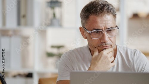 Casual mid adult man with laptop computer in home office, banking online, remote working. Portrait of older bearded guy thinking. Businessman, entrepreneur managing business on internet.