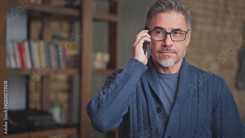 Middle aged man on phone call at home. Gray haired older male in glasses and sweater talking on mobile in living room. Telecommunication .