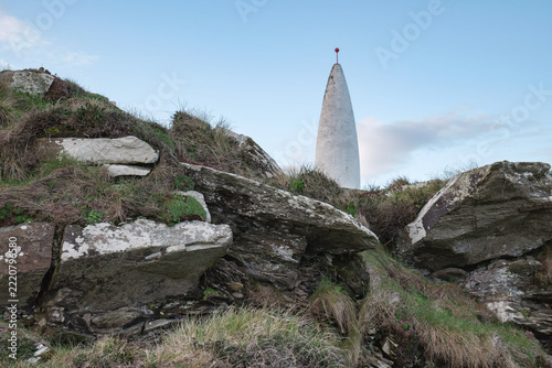 A tall, conical white structure with a red spherical top stands against a pale blue sky, partially obscured by rocky terrain.