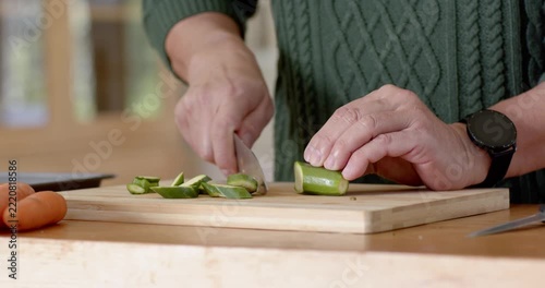 Hands are positioning halved cucumber then slicing thin rounds on wooden board for salad