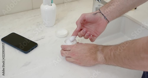 Hands and arms are reaching opening jar scooping cream and rubbing palms hydrating skin near sink