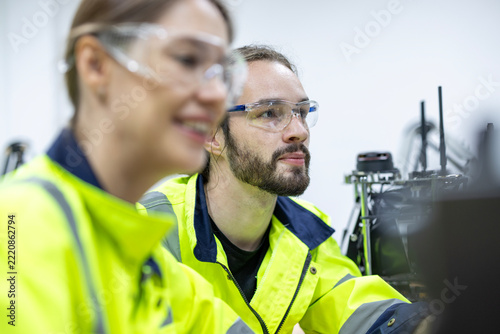Robotics engineers in high-visibility jackets calibrating robotic arm machine. Industrial technicians collaborating on desktop automation prototype in high-tech lab. Male and female tech professionals