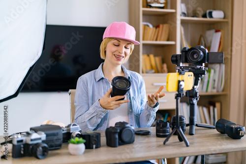 A caucasian woman in a pink cap is reviewing camera equipment for her online audience, surrounded by photography gear