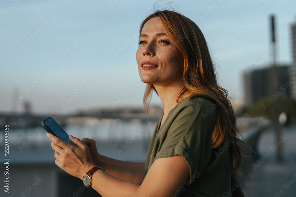 Naklejka premium Woman holding smartphone reflecting at sunset cityscape