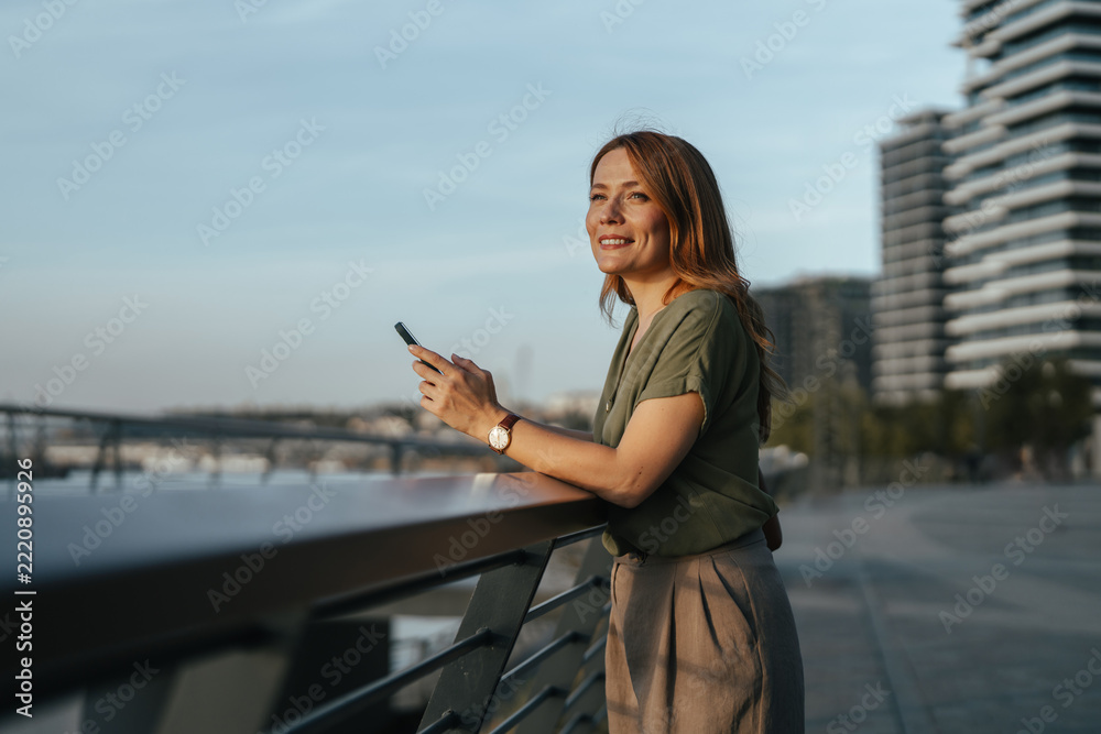 Naklejka premium Woman smiling holding smartphone watching urban cityscape