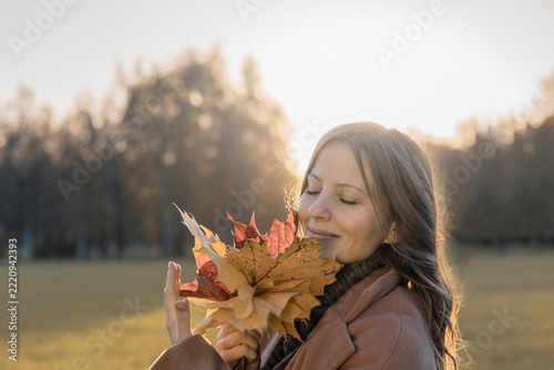 Young woman with closed eyes smiling gently while holding colorful maple leaves near her face at autumn sunset, peaceful fall lifestyle with copy space