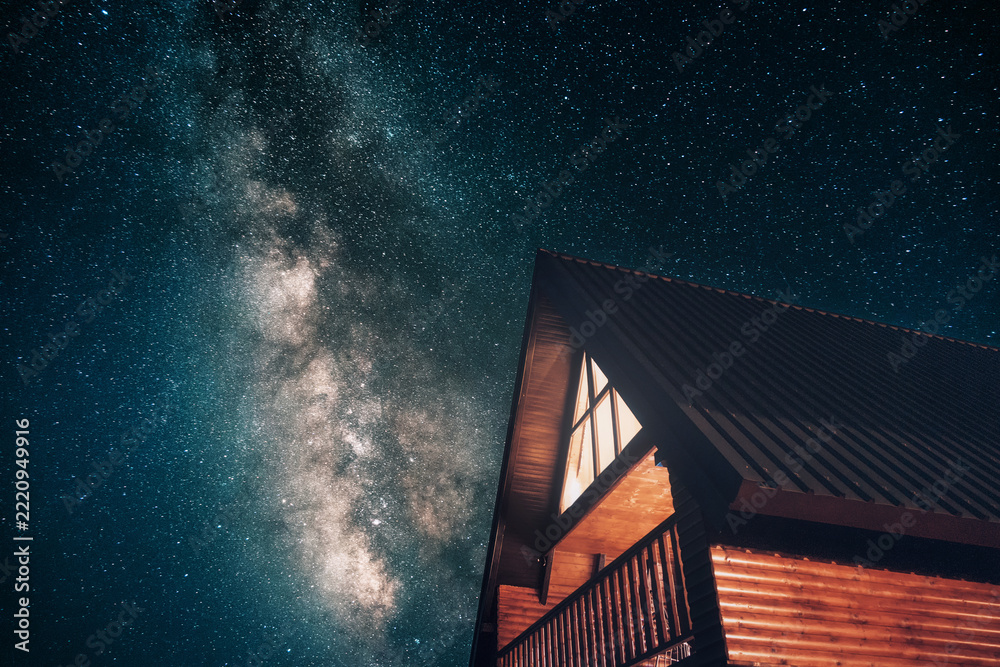 Fototapeta premium Wooden cabin illuminated at night, standing out against a dark sky filled with countless stars and the visible Milky Way galaxy
