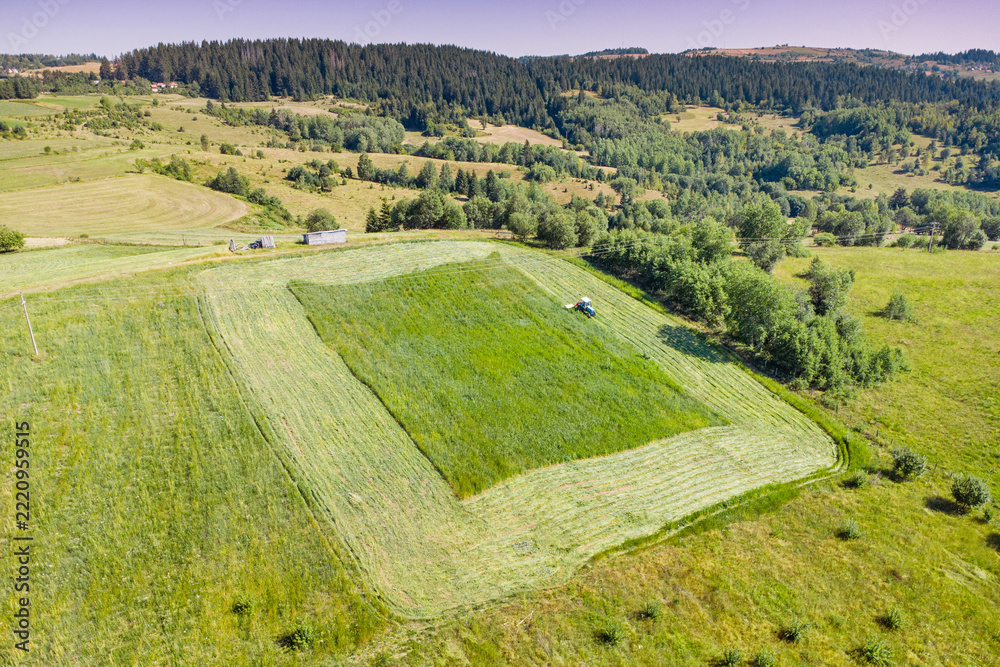 Naklejka premium Aerial view of a tractor mowing green meadow fields on a sunny summer day - ideal for agricultural and rural lifestyle concepts