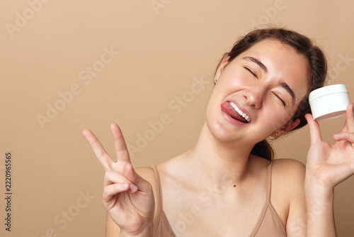 Fotografie Playful youth lifestyle, funny young woman sticks out her tongue, shows peace sign and holds white cream jar on beige background