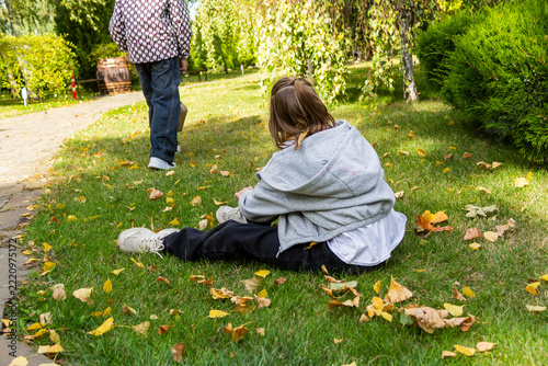 fair-haired girl sits by path on green grass among autumn leaves in an autumn park, resting, bright yellow leaves, silence, in fall, child, picturesque background,