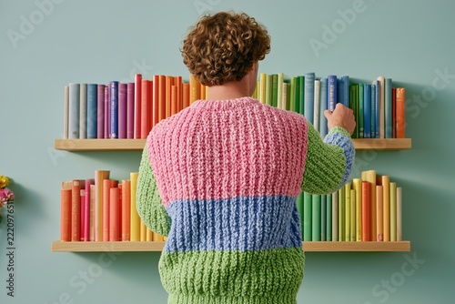 Young man in colorful knitted sweater arranging rainbow books on minimalist wooden shelves