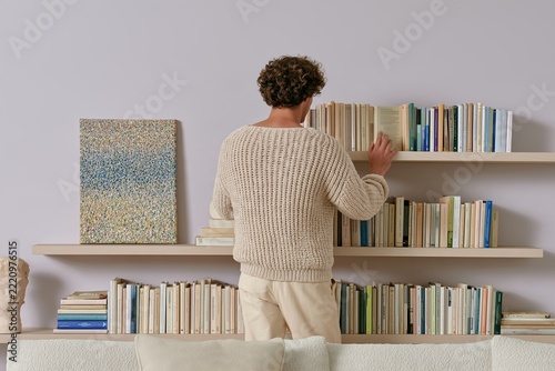 Young man organizing books on minimalist wooden bookshelves in bright contemporary living room