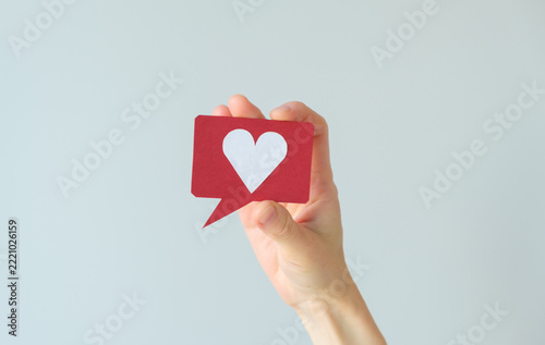Social media and digital communication concept photo featuring a hand holding a red speech bubble icon with a white heart symbol on a light background, representing likes.