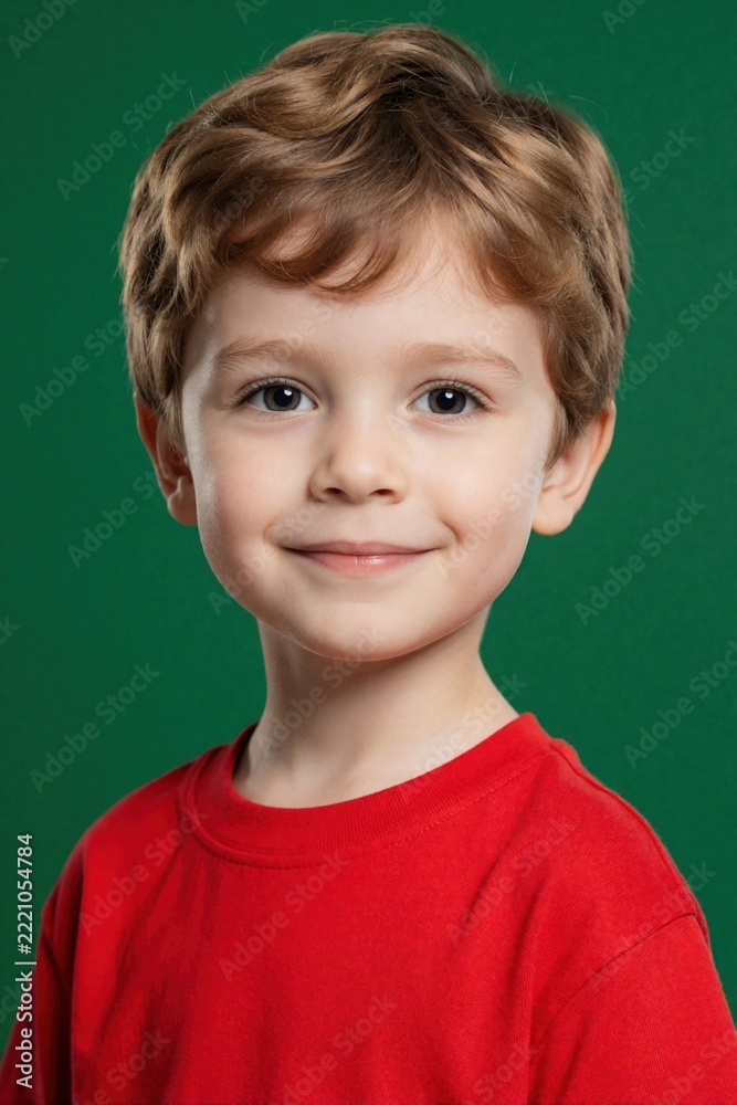 Fototapeta premium Cute little boy with blond hair wears red t-shirt. Smiling young kid looks at camera in studio. Portrait of happy child with bright eyes on green background. Childhood happiness, innocence concept