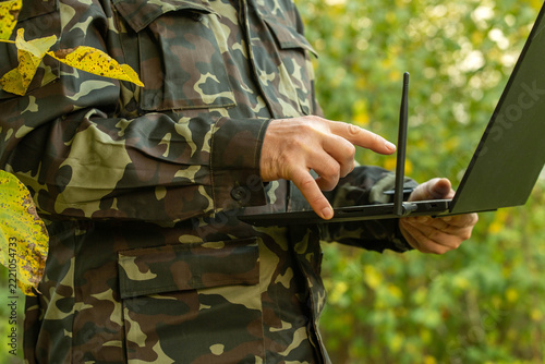 Soldier in camouflage presses laptop keys while holding computer outdoors, focused on its screen. Field operator uses portable equipment with antenna amid leafy greenery for tactical communications.