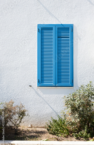 Blue louvered window shutter on textured white exterior wall with sunlight, shadows, and surrounding shrubs in a Mediterranean architectural detail
