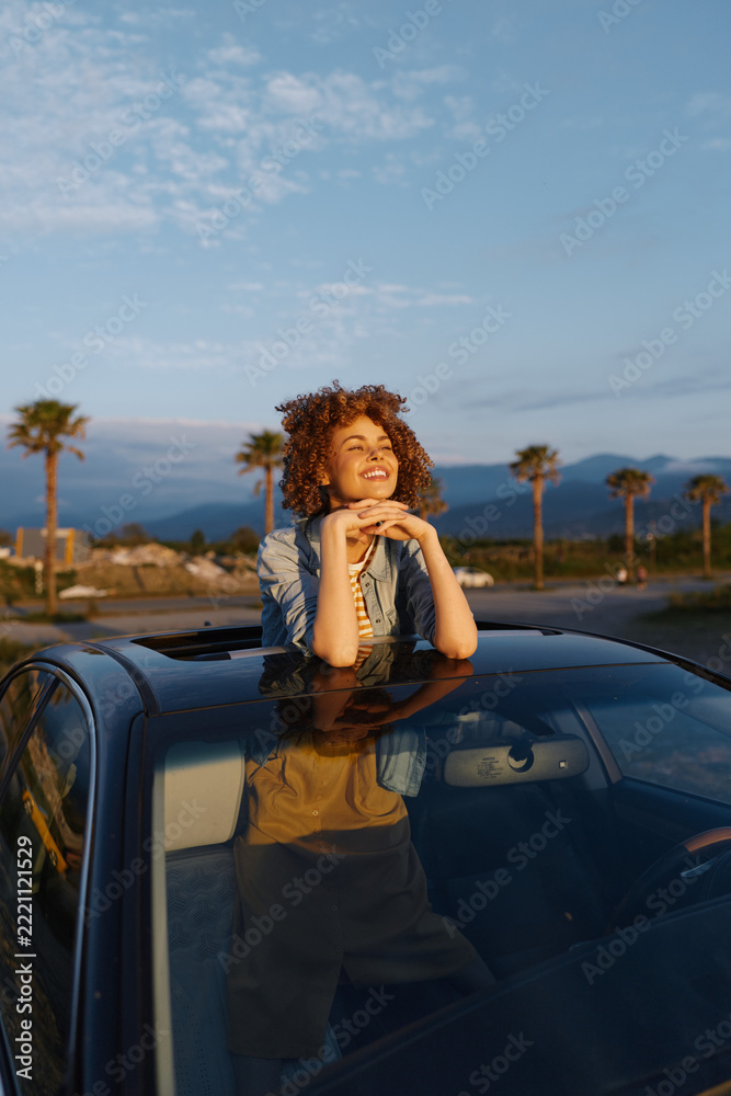 Fototapeta premium Casual woman leaning on car hood, smiling with curly hair, sunny outdoor scene, desert landscape with palm trees, relaxed lifestyle, warm glow, carefree mood, candid moment in bright light.