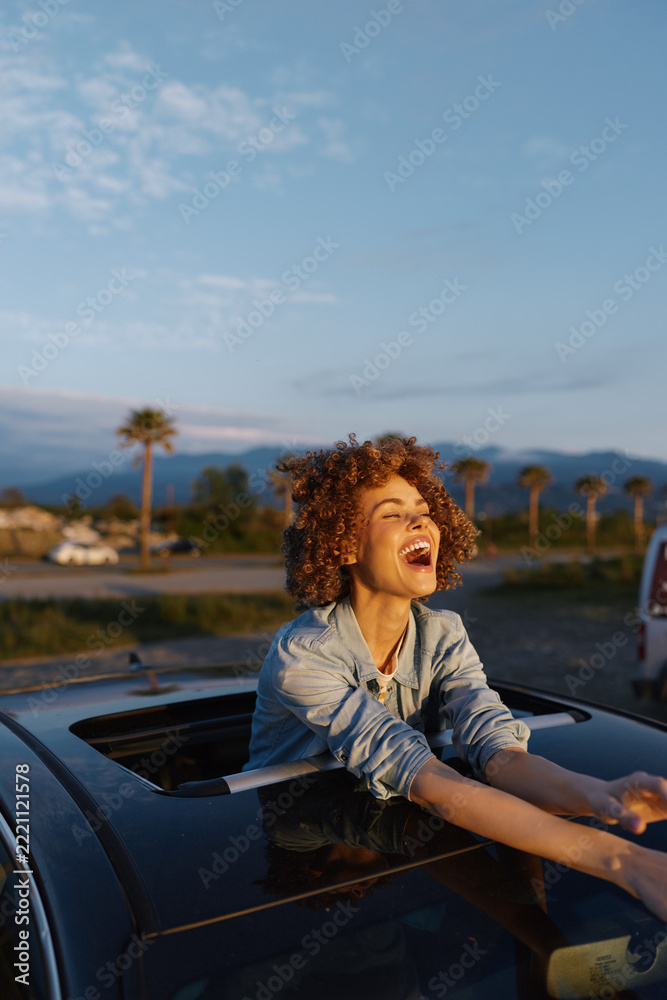 Fototapeta premium Young woman leaning out of a car window, laughing with curly brown hair in a denim jacket, sunset light and palm trees in the background, mountains on the horizon.