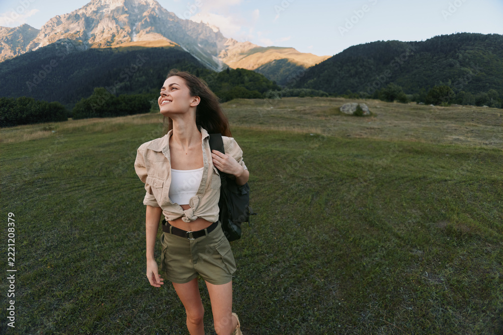 Naklejka premium Young woman enjoys a sunny hike in a grassy field with distant mountains, backpack slung over one shoulder as she gazes upward with a smile.