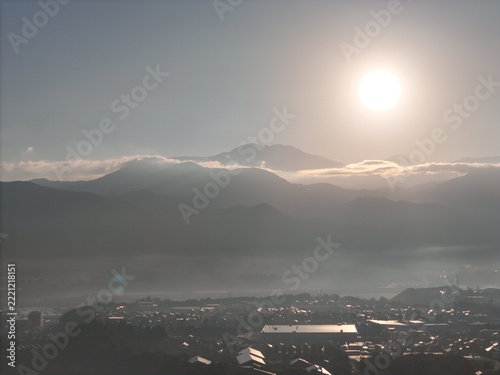 A wide warm sky, high bright sun and thin cloud banks frame dark mountain layers and a hazy valley town.