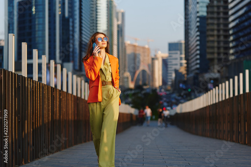 Strategy consultant, confident young woman in orange blazer and sunglasses talking on smartphone walking across city bridge with skyscrapers. Concept of business success, urban lifestyle and ambition.