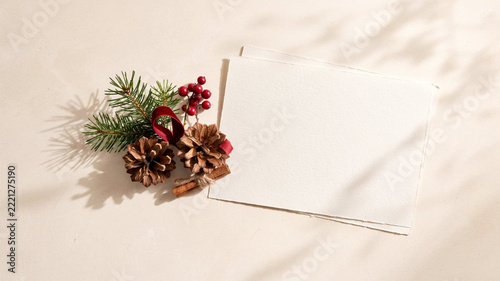 Minimalist Christmas Greeting Card Mockup with Blank Textured Paper, Pine Cone, Fresh Evergreen Sprig, Red Winterberries and Velvet Ribbon on Warm Cream Surface
