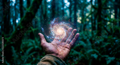 A human hand holding a glowing spiral galaxy surrounded by sparkling stars in a mysterious forest. Surreal cosmic energy blends with lush green woodland