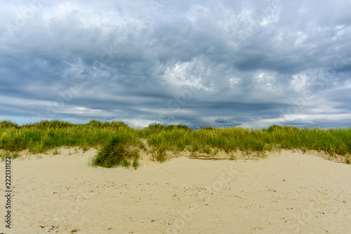 Sandy beach dunes creating a natural barrier along the coast, topped with lush green marram grass, all beneath a dramatic, overcast sky suggesting approaching weather